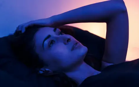 Młoda kobieta leży w łóżku, ręka oparta o głowę. Nocne światło tworzy intymną atmosferę, idealną na seks po kryjomu.