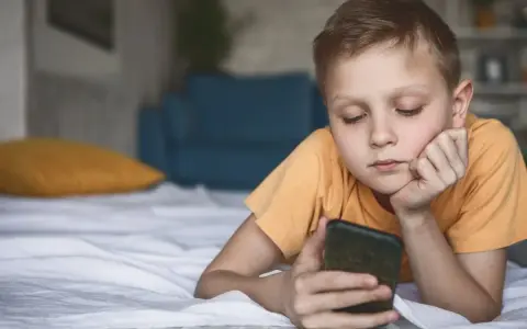 Chłopiec leży na łóżku, patrząc na telefon. Zastanawia się, co to jest sexting i jakie niesie zagrożenia.