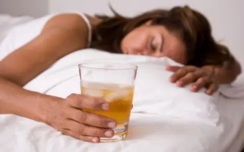 Kobieta w łóżku z drinkiem, nieświadoma. Objawy po zażyciu pigulka gwaltu mogą być różne.