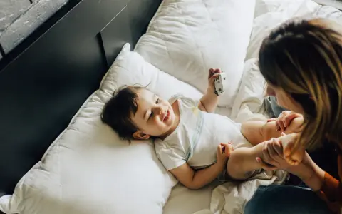Uśmiechnięte dziecko bawi się zabawką na łóżku. Mama patrzy na malucha. Co to za mastka?