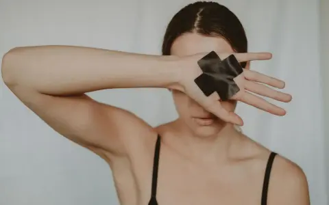 Kobieta zakrywa oczy dłonią z czarną taśmą w kształcie krzyża, symbolizującą trauma seksualna i potrzebę uzdrowienia.